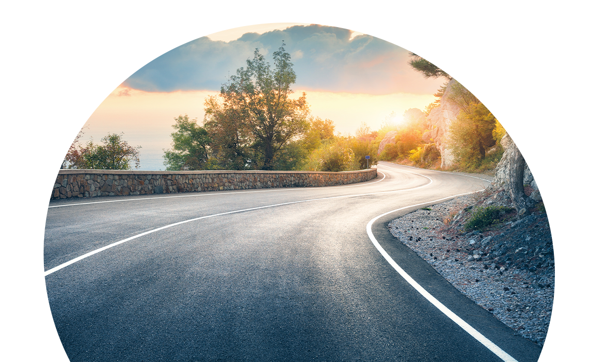 Winding coastal road at sunset, curves around a stone guardrail with trees in the background.