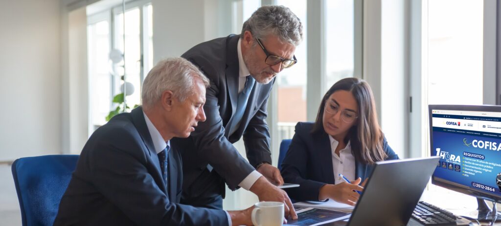 Three professionals in a meeting around a desk with laptops, a man points at a tablet as a colleague writes notes.