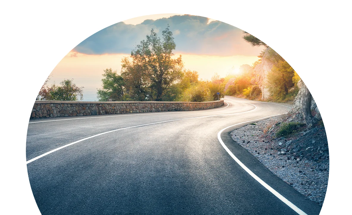 Winding coastal road at sunset, curves around a stone guardrail with trees in the background.
